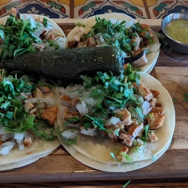 Four chicken tacos with cilantro, onions, a roasted jalapeño, lime, and green sauce on a wooden board.