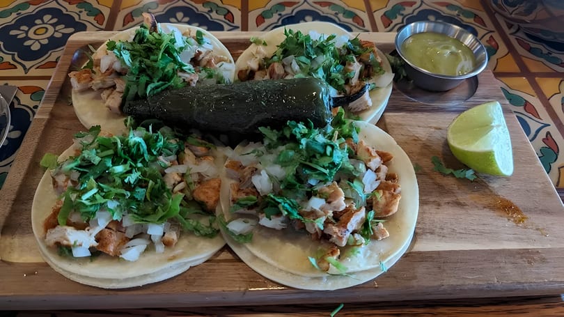 Four chicken tacos with cilantro, onions, a roasted jalapeño, lime, and green sauce on a wooden board.