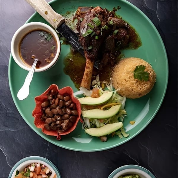 Braised lamb shank with rice, beans, salad, and avocado on a green plate with sides of salsa and guacamole.