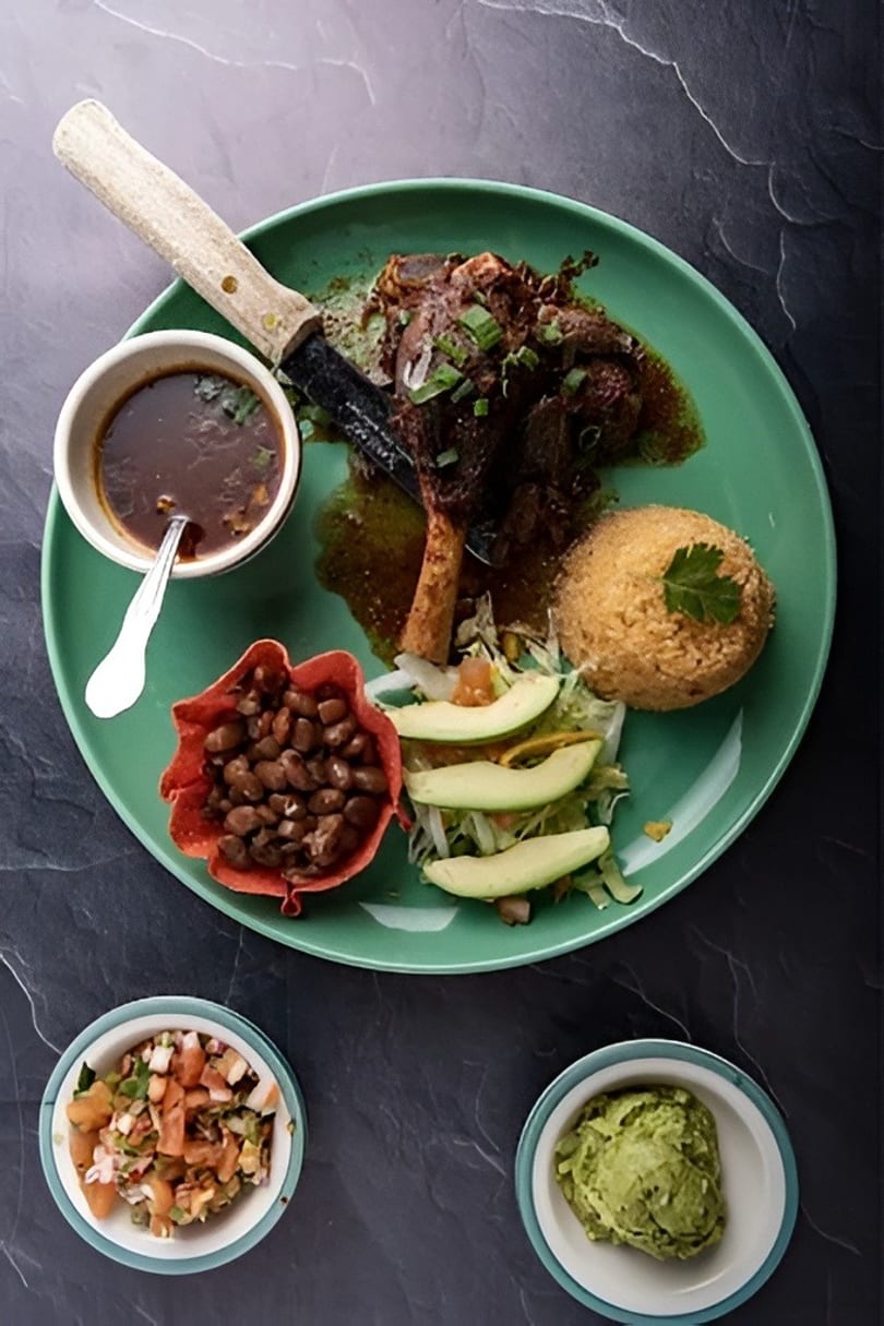 Braised lamb shank with rice, beans, salad, and avocado on a green plate with sides of salsa and guacamole.