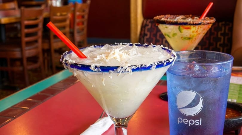 Two margaritas, one coconut and one spicy, with a Pepsi on a red table in a restaurant.