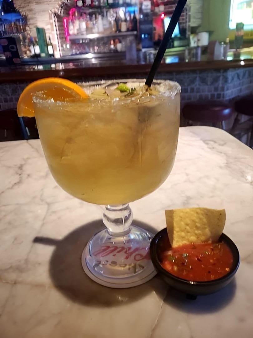 Margarita with orange slice and straw next to a bowl of salsa and a tortilla chip.