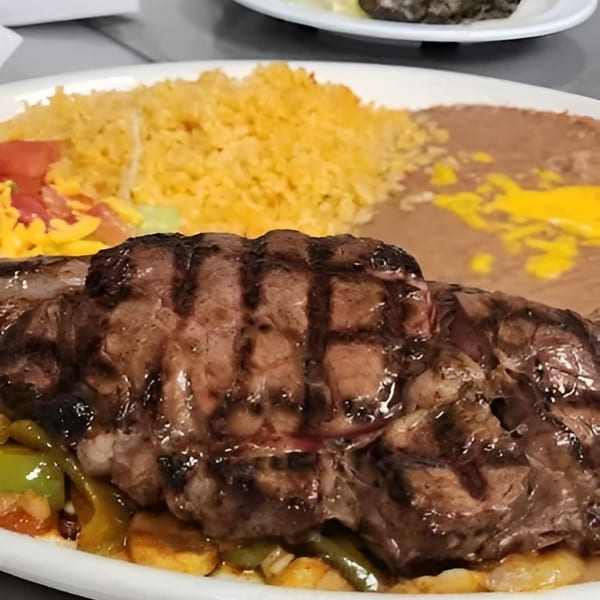 Grilled steak with rice, refried beans, and vegetables on a white plate, with a side salad.