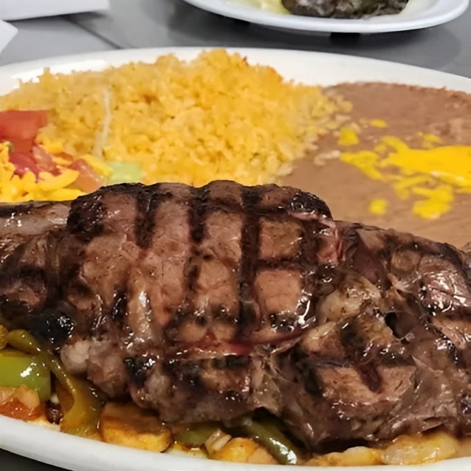 Grilled steak with rice, refried beans, and vegetables on a white plate, with a side salad.