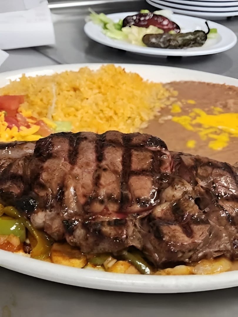 Grilled steak with rice, refried beans, and vegetables on a white plate, with a side salad.